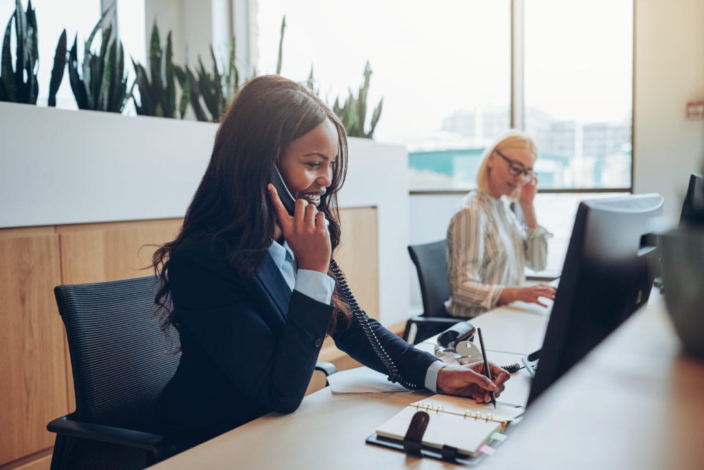 Smiling young African American businesswoman talking on a telephone while working with a colleague at an office reception desk