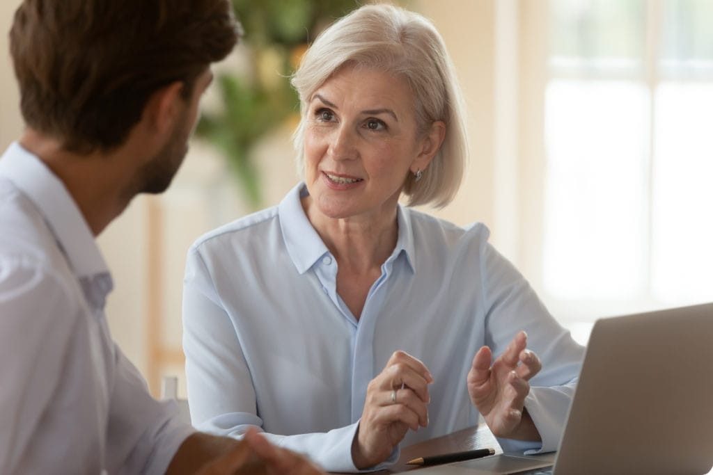 Middle aged businesswoman manager mentor speaking to businessman client teaching intern or convincing customer at business office meeting sit at table.