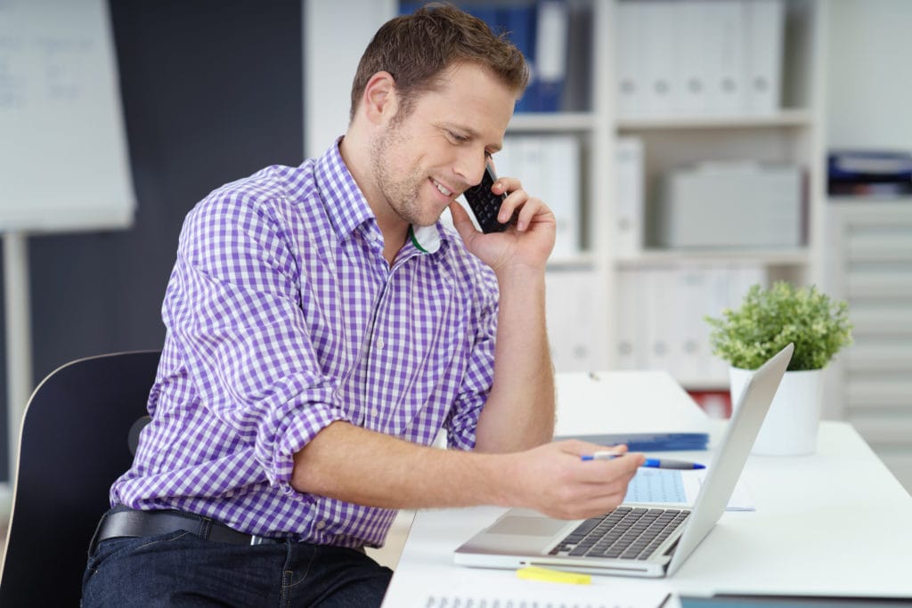 man sitting at white desk in front of laptop holding cell phone to ear