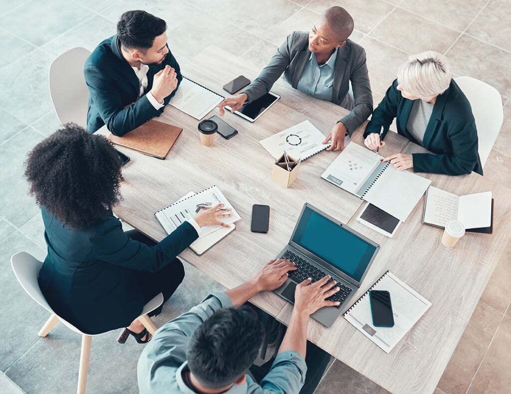 Sales team sitting around table working on laptops with cell phones and paperwork scattered around the table with the focus on Lead generation trends 2025