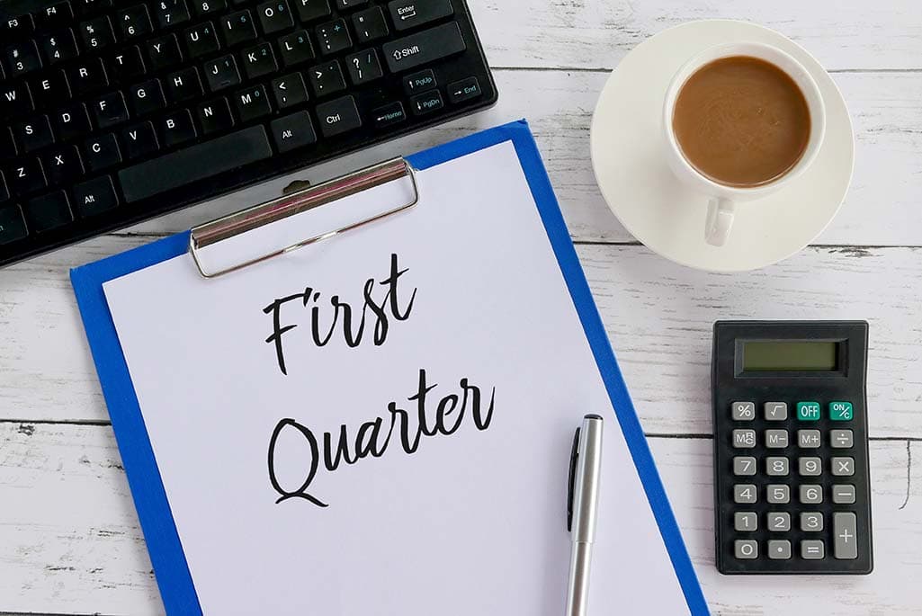 Computer keypad next to a note pad with First Quarter written on it next to a coffee and calculator focusing on Q1 success metrics.